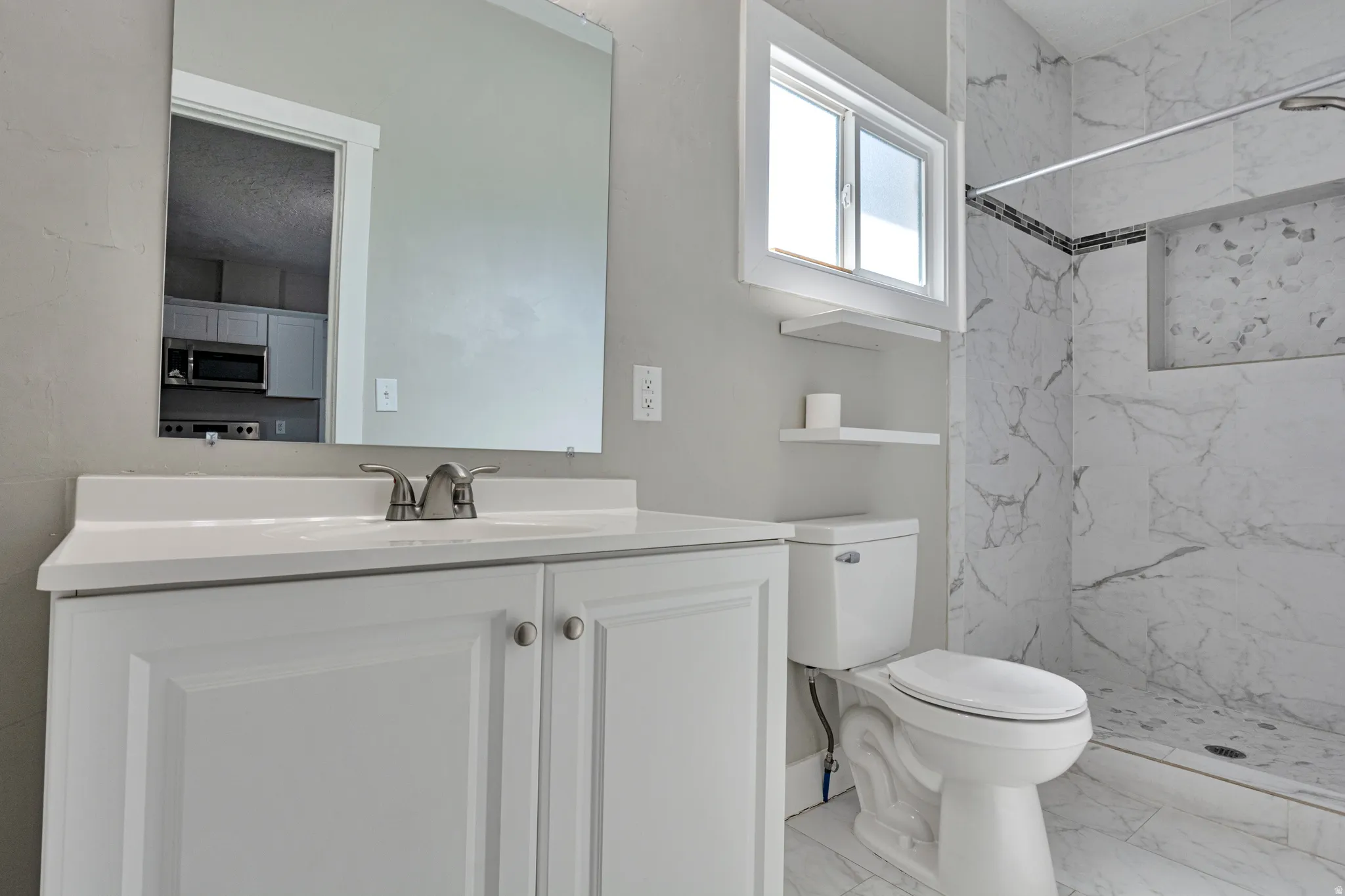 Bathroom featuring a white vanity with brushed nickel hardware, a rectangular vanity mirror, a window with a sill, and a tiled shower with a built-in niche