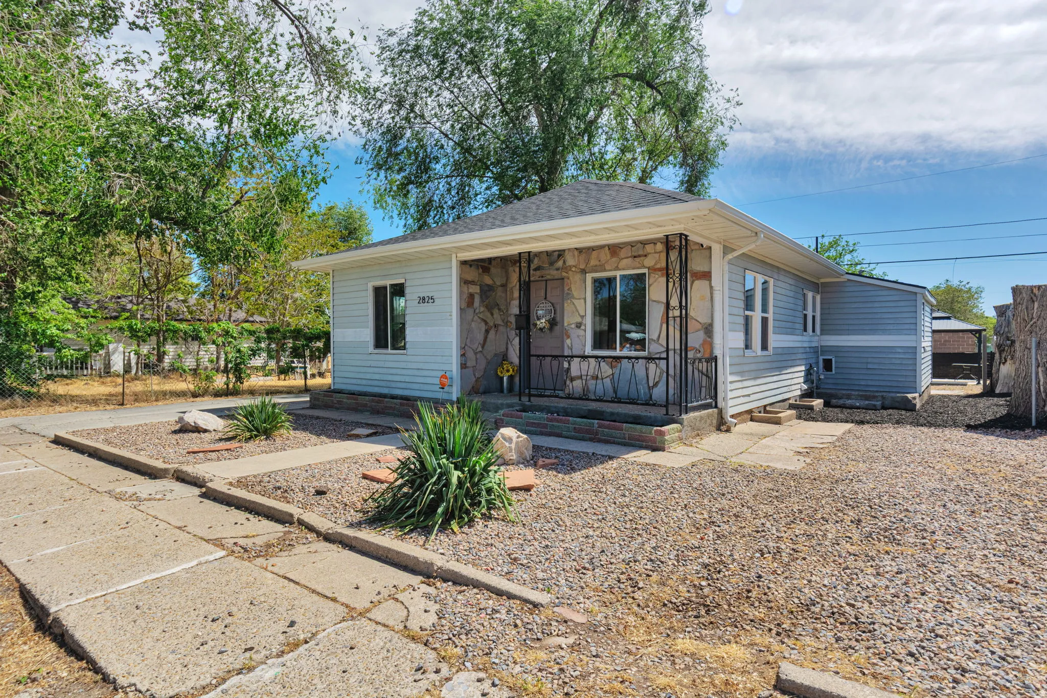 Exterior featuring light blue siding with stone veneer accents, a covered front porch with wrought iron railing, a gravel front yard, mature trees, and a concrete pathway