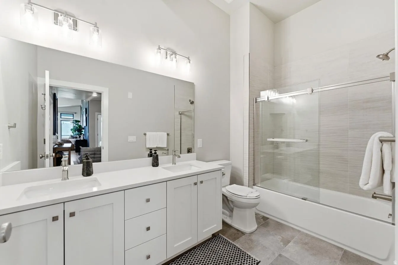 Bathroom featuring a dual-sink vanity with white cabinetry and a light-toned countertop, a large wall-mounted mirror, a bathtub with a sliding glass door, and tiled flooring