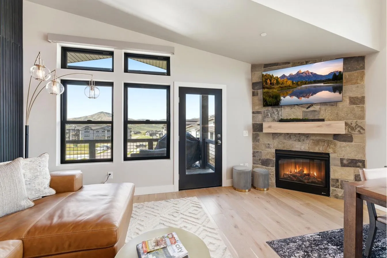 Living area featuring wide plank wood-finish flooring, a stone-clad fireplace with a wood mantel, a glass-paned exterior door, and multiple windows with black frames