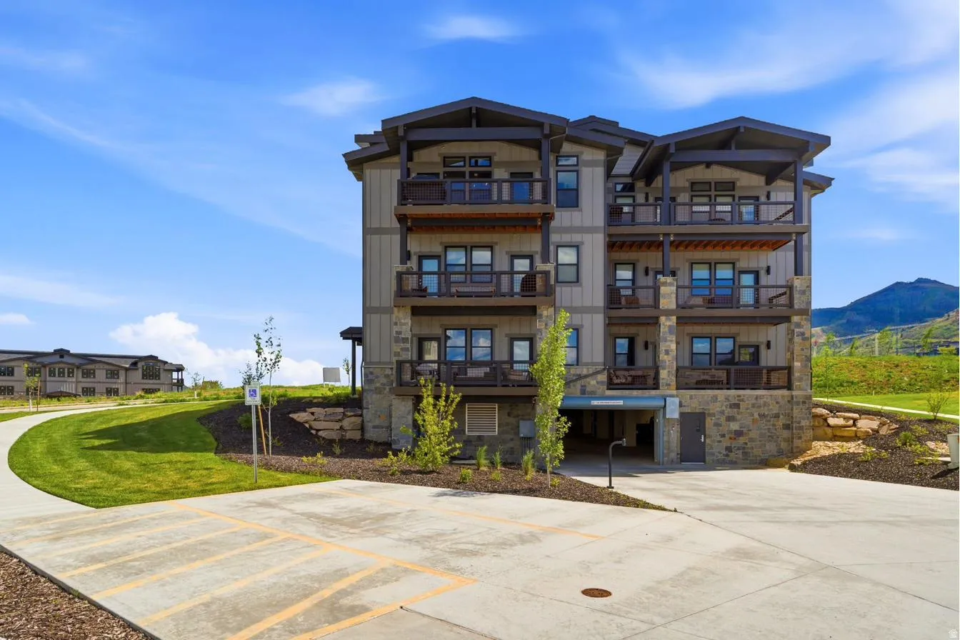 Multi-story residential building featuring stone and siding exterior construction, multiple private balconies with dark railings, a ground-level covered parking area, and landscaped grounds with a paved pathway
