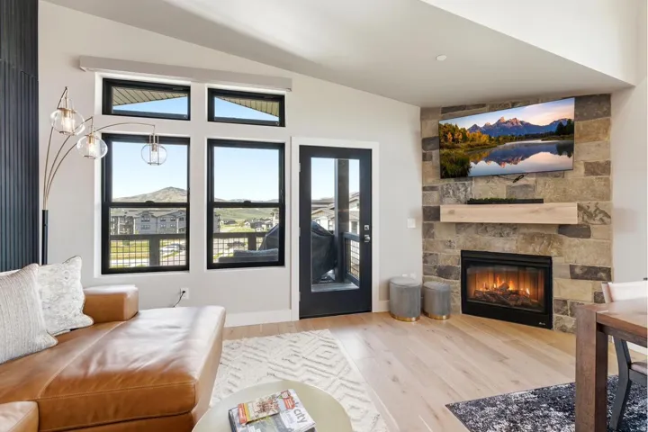 Living area featuring wide plank wood-finish flooring, a stone-clad fireplace with a wood mantel, a glass-paned exterior door, and multiple windows with black frames