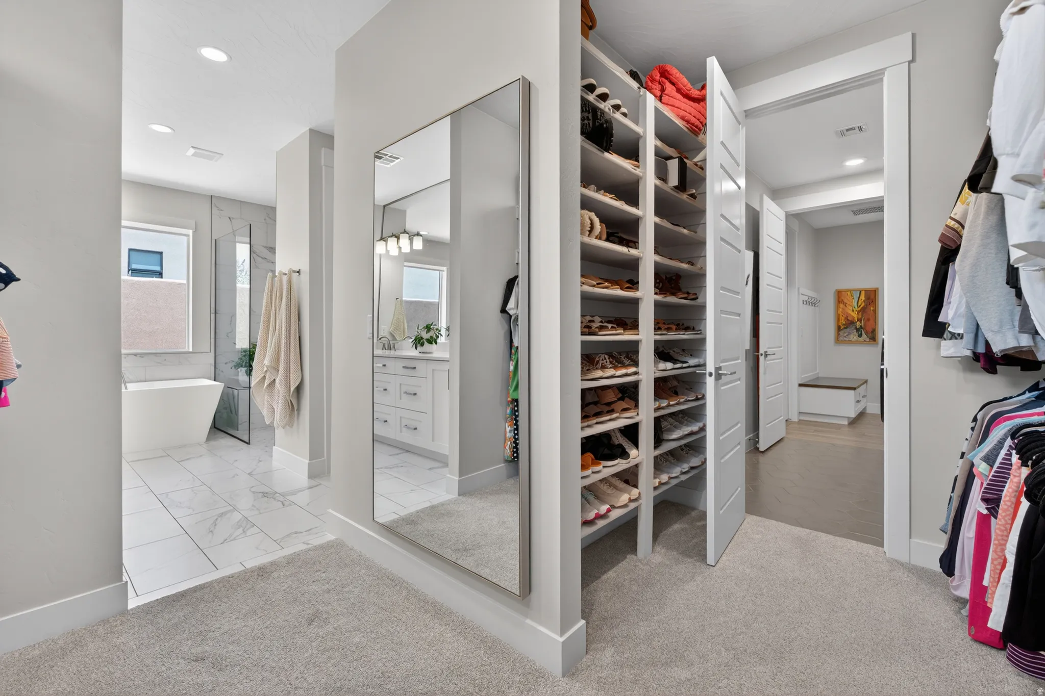 Walk-in closet featuring extensive built-in shoe storage, full-length wall mirror, light-colored carpeting, and direct access to a bathroom with a freestanding tub and walk-in shower
