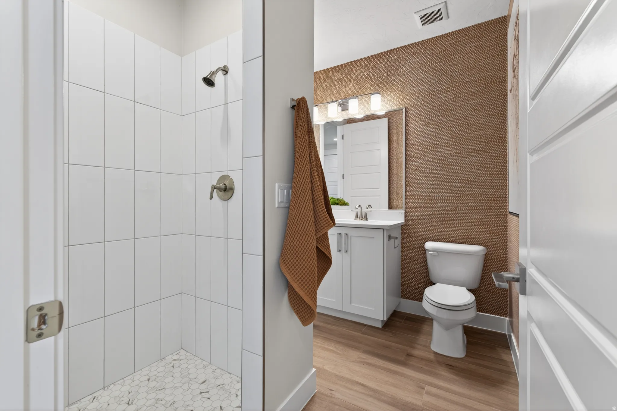 Bathroom featuring a walk-in shower with vertical subway tile, hexagon tile flooring, a single vanity with a white countertop, wood-finish flooring, and textured wall covering