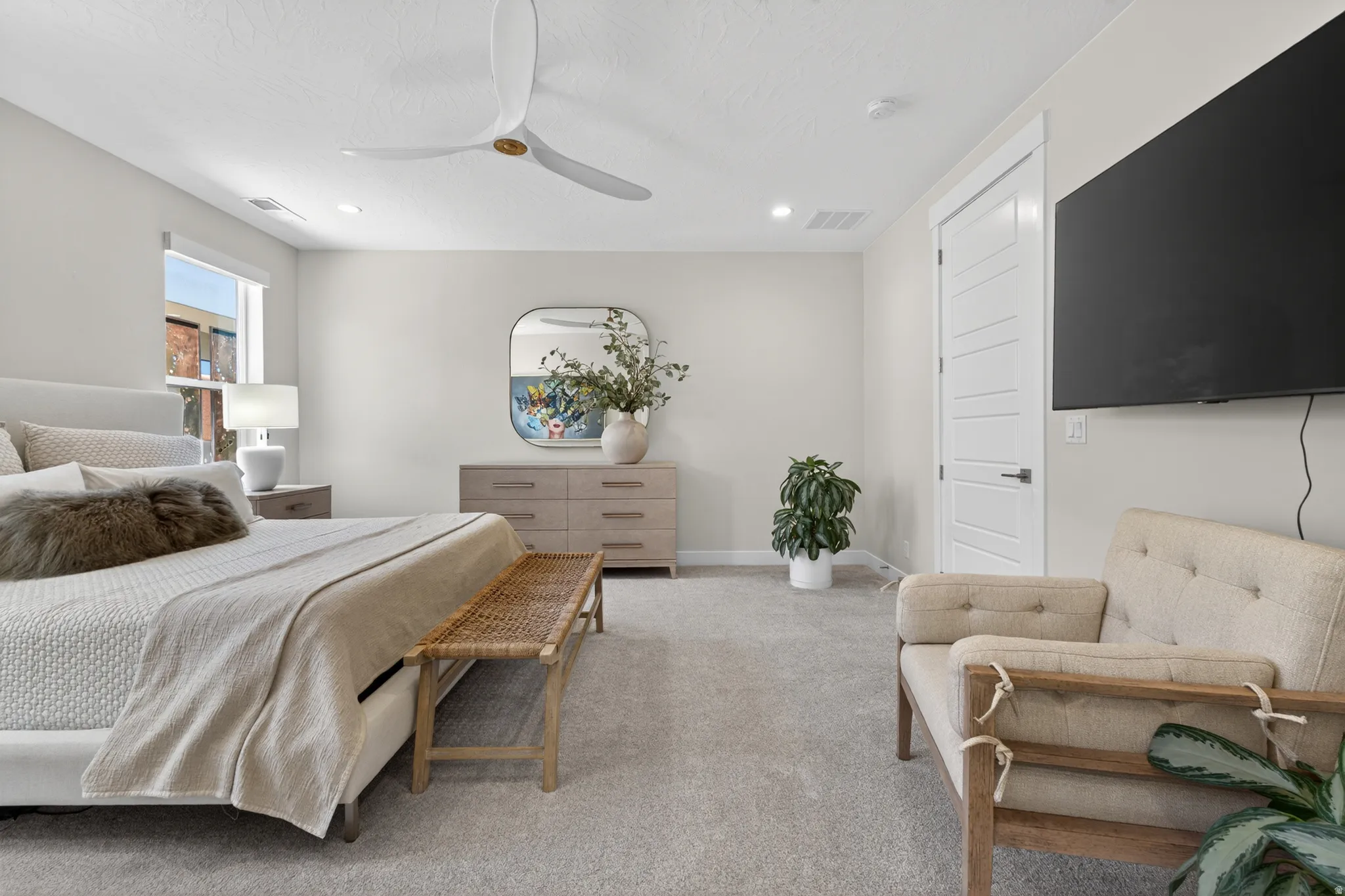 Spacious bedroom featuring light neutral wall tones, light gray carpeting, a large window with natural light, a modern ceiling fan, and a paneled interior door