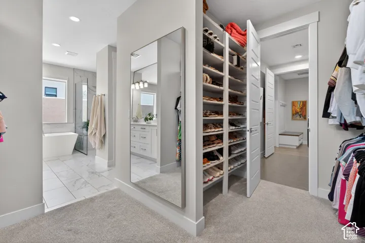 Walk-in closet featuring extensive built-in shoe storage, full-length wall mirror, light-colored carpeting, and direct access to a bathroom with a freestanding tub and walk-in shower