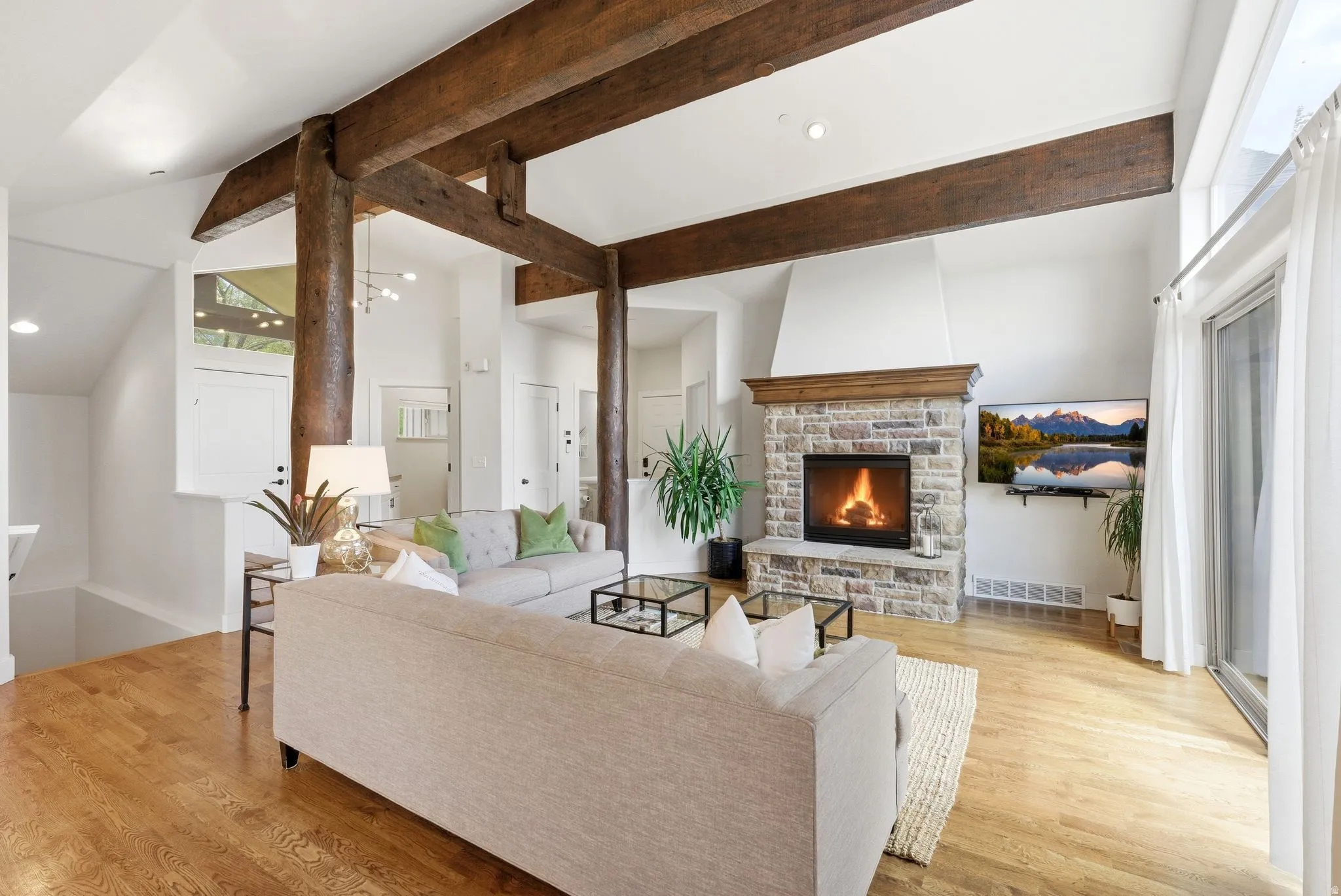 Spacious living area featuring exposed wood beams, a vaulted ceiling, wood-finish flooring, a stone fireplace with a wood mantel, and sliding glass doors