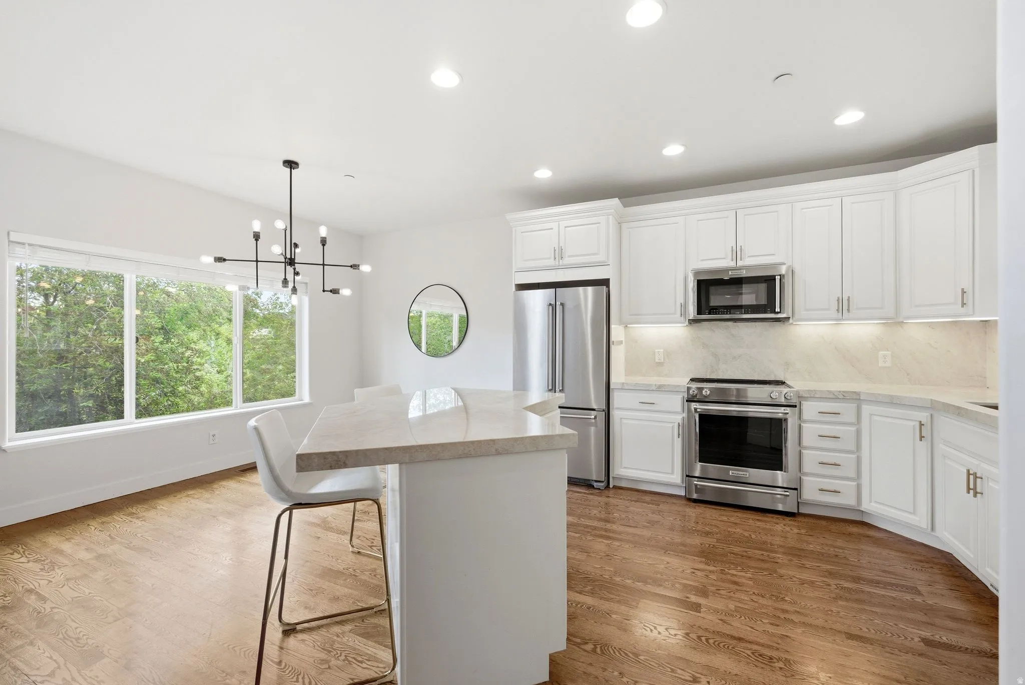 Open-concept kitchen and dining area featuring hardwood flooring, white cabinetry, stainless steel appliances, a light-colored countertop with an integrated island, and a contemporary chandelier