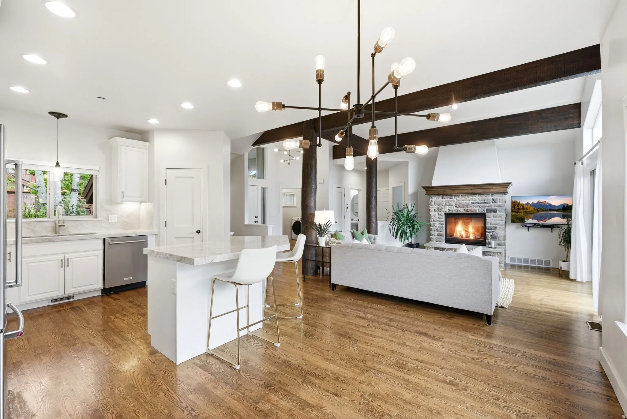 Open-concept living area featuring wood-finish flooring throughout, a stone-front fireplace with a wood mantle, and exposed ceiling beams