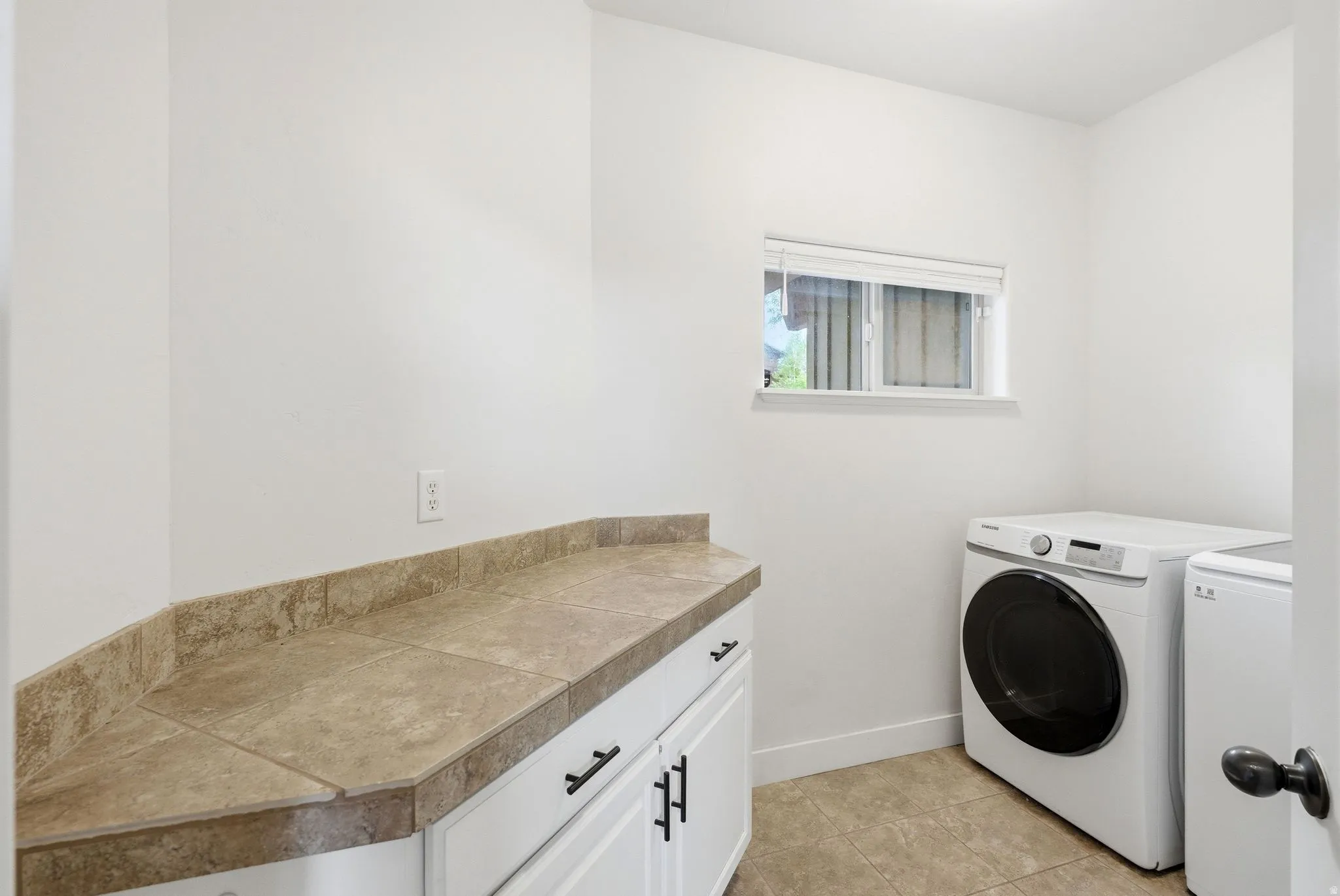 Functional laundry space featuring white cabinetry with black hardware, a tiled countertop, and a window