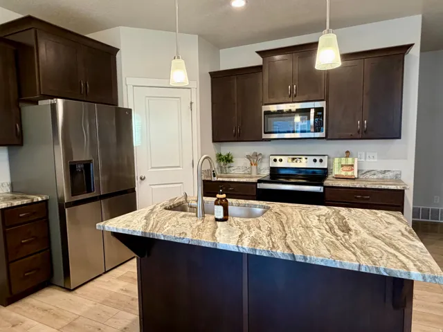 Kitchen featuring a central island with an undermount sink, dark wood-finish cabinetry, stainless steel appliances, pendant lighting, and wood-look tile flooring
