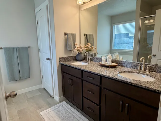 Dual vanity with granite countertop and integrated sinks, brushed nickel fixtures, dark wood-finish cabinetry, a large wall-mounted mirror, and tile flooring