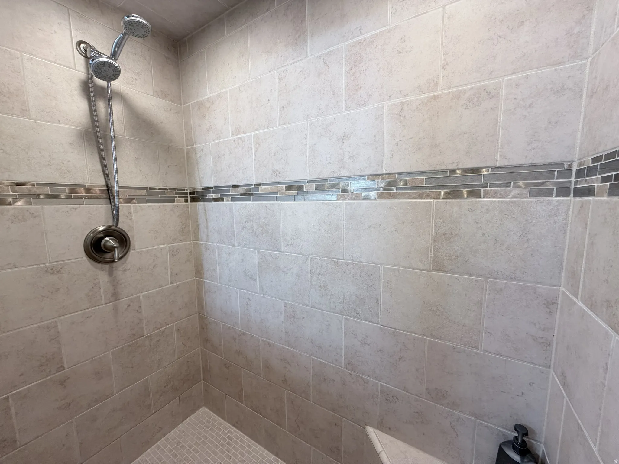 Shower stall featuring large format rectangular wall tiles, a decorative accent band of mosaic tiles, a handheld shower fixture, and a built-in corner bench