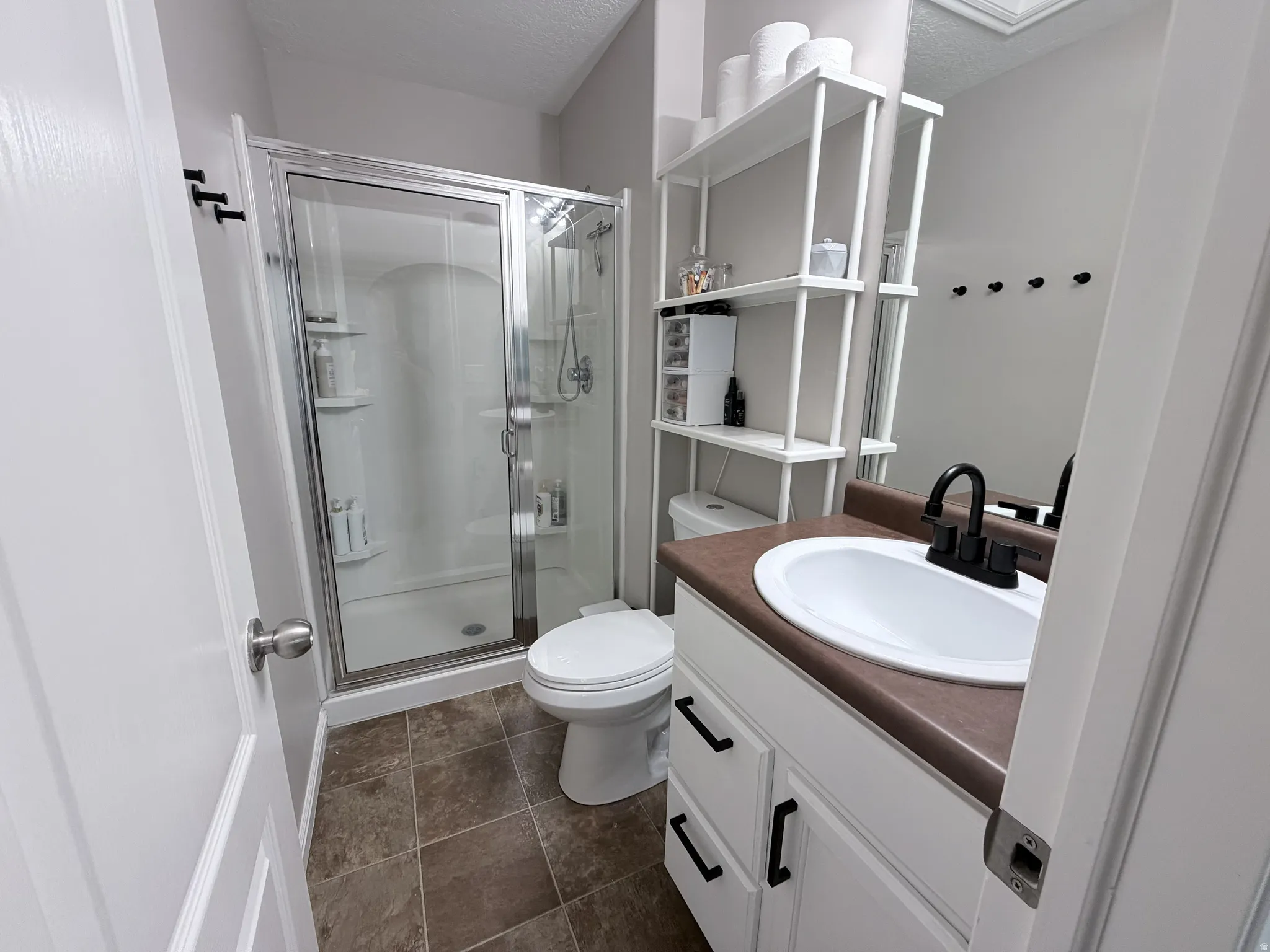 Shower enclosure with clear glass doors, white vanity with integrated sink, dark countertop, and matte black faucet, complemented by brown tile flooring