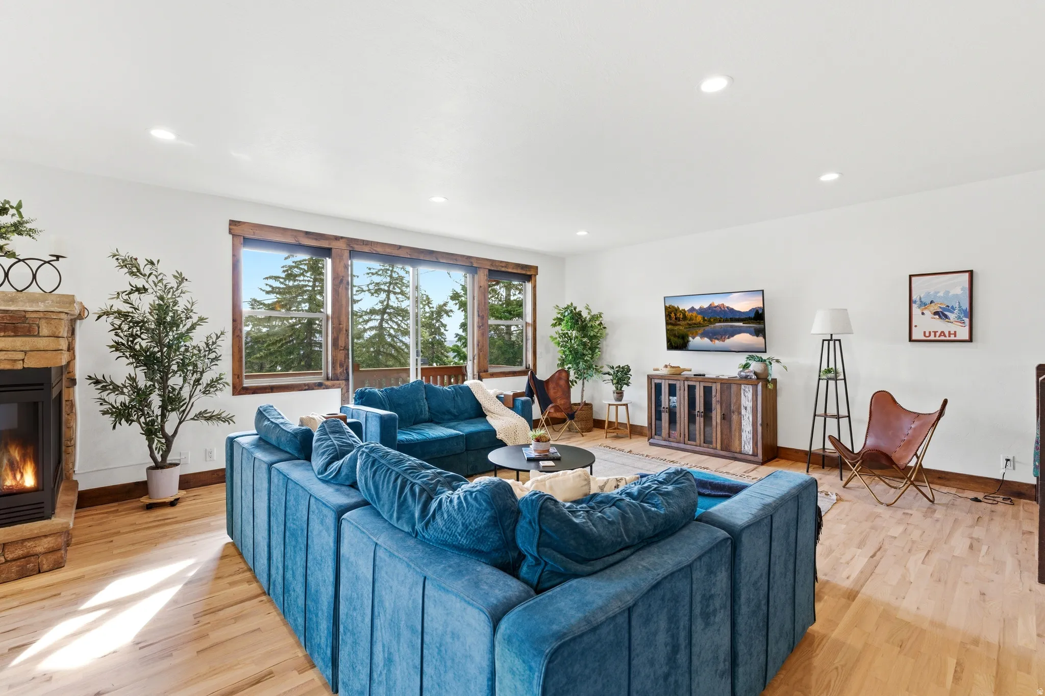 Spacious living area featuring light wood-finish flooring, a stone-clad fireplace, and large windows with natural wood trim