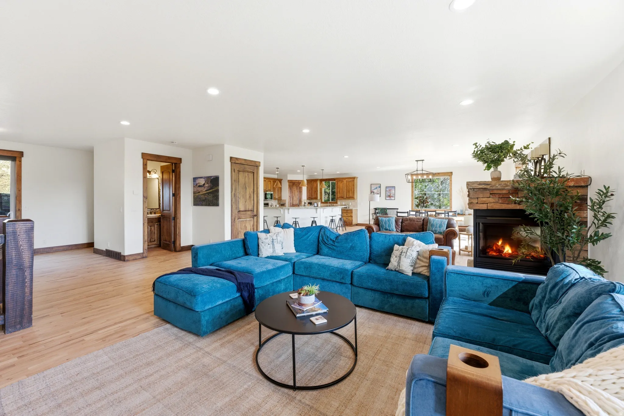 Spacious living area featuring light wood-finish flooring, a stone-clad fireplace, recessed lighting, and a visible kitchen space with wood cabinetry