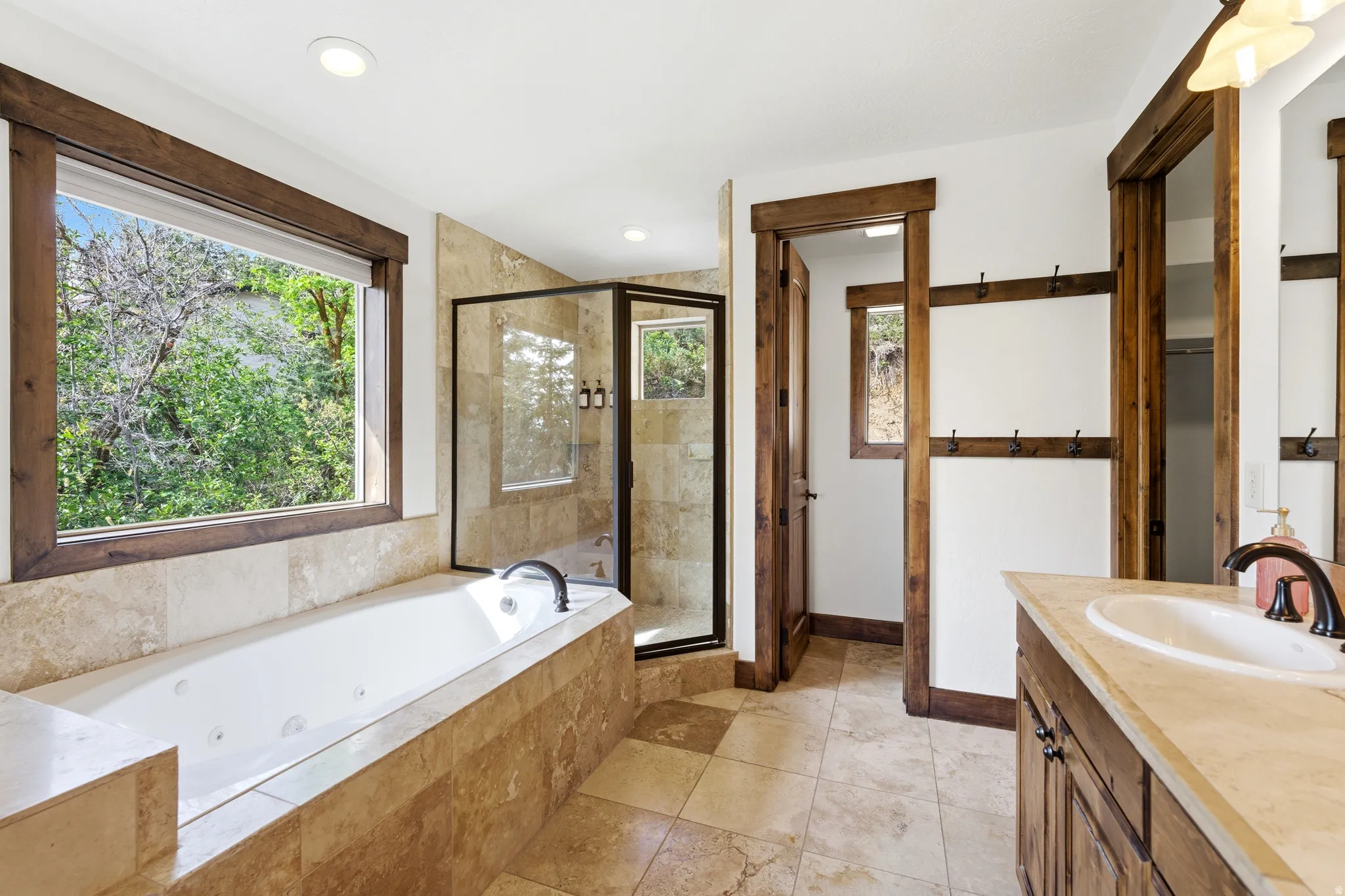 Spacious bathroom featuring a built-in jetted tub, a glass-enclosed shower, stone-finish tile flooring, a single vanity with a stone countertop, and wood-finish trim and cabinetry