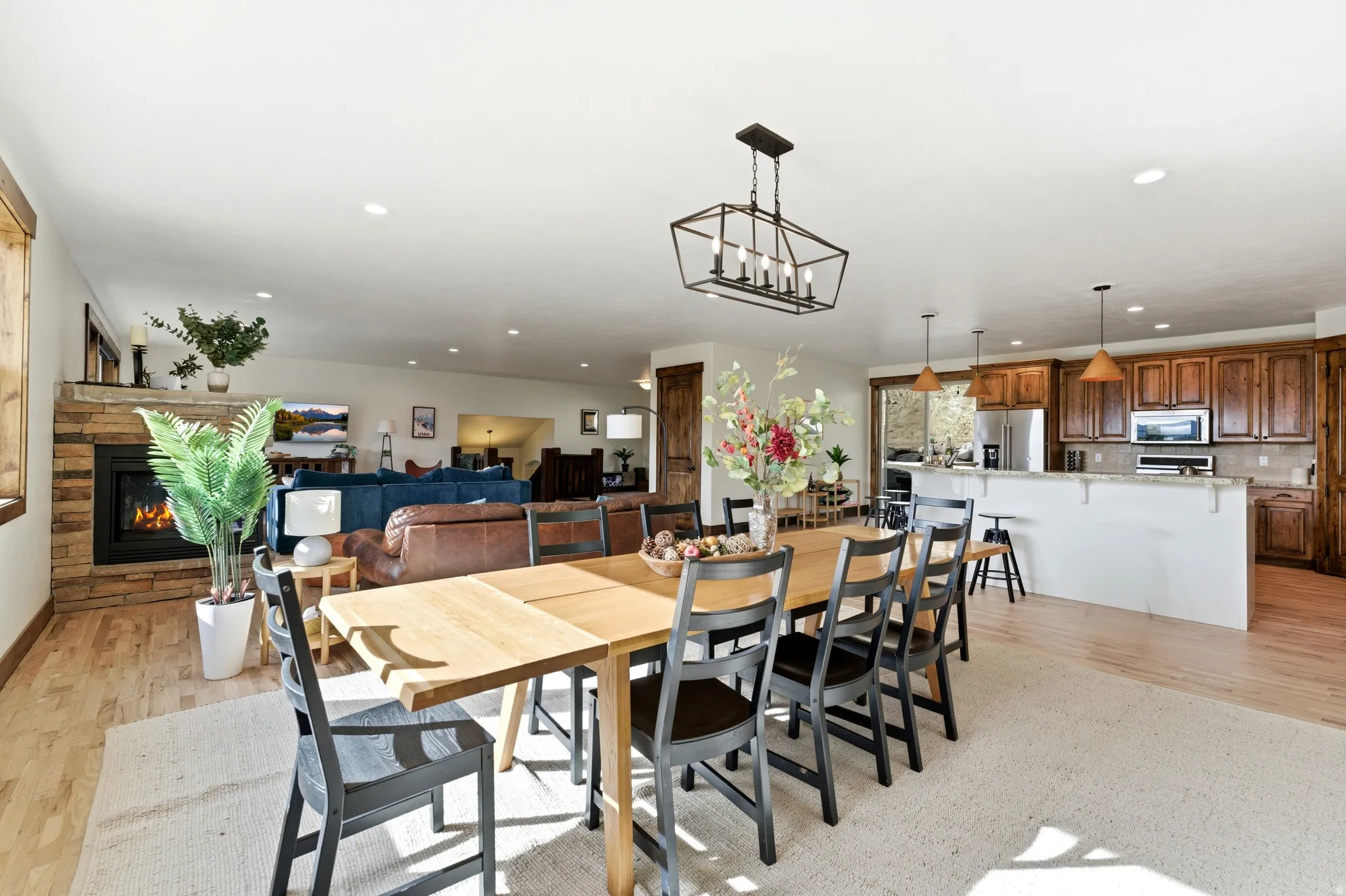 Open-concept living space featuring a dining area with a modern linear chandelier, wood-finish flooring, a stacked stone fireplace, and a kitchen with warm wood cabinetry and stainless steel appliances