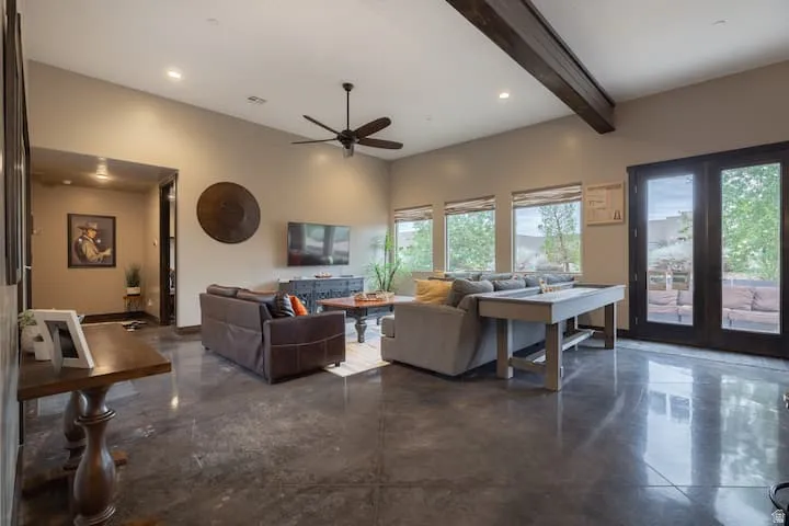 Spacious living area featuring polished concrete flooring, exposed ceiling beams, multiple windows, recessed lighting, and dark-framed double doors leading to an outdoor space
