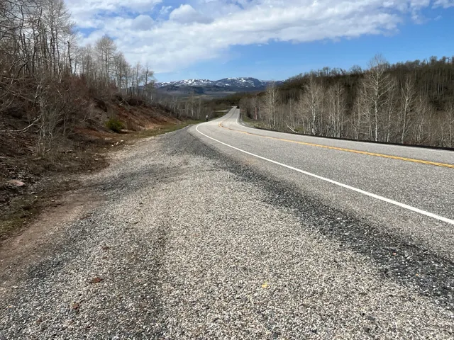 Paved roadway with a gravel shoulder, flanked by wooded hillsides featuring deciduous trees