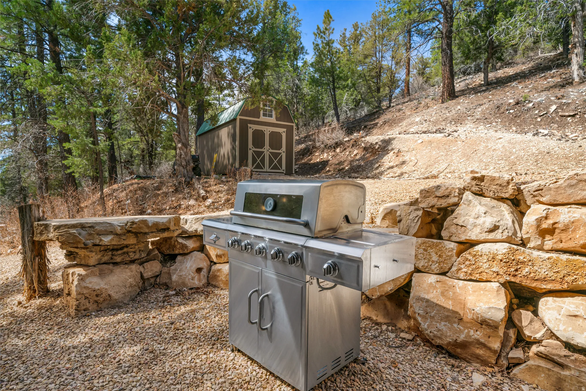 Outdoor stone retaining wall and seating area on a gravel patio, featuring a built-in storage shed and mature trees