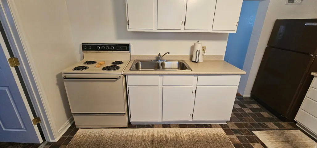 Compact kitchen featuring white upper and lower cabinetry, a double basin stainless steel sink, a beige countertop, and brown patterned floor tiles