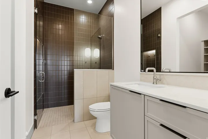 Bathroom featuring a walk-in shower with vertical tile surround and glass door, a single vanity with a white countertop and undermount sink, and light-toned floor tiles