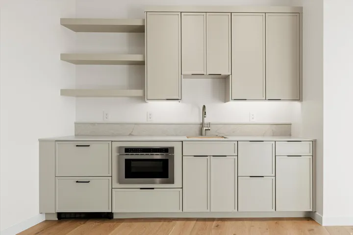Built-in wet bar featuring light wood-finish cabinetry, stainless steel sink with gooseneck faucet, integrated microwave, stone-finish countertops with matching backsplash, and three floating shelves
