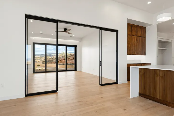 Expansive glass sliding doors open to desert scenery, complemented by light wood-finish flooring, a contemporary ceiling fan, recessed lighting, and rich wood cabinetry with integrated countertops
