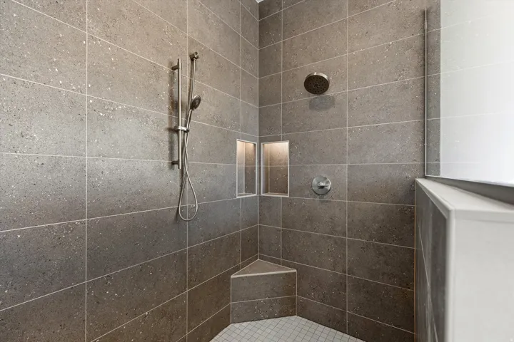 Shower featuring large format gray speckled wall tiles, rain shower head, handheld shower fixture, built-in corner bench, and integrated niche shelving