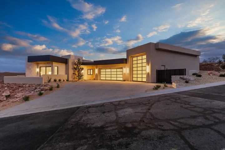 Contemporary desert architecture featuring a stucco exterior, multiple garage doors with frosted glass panels, an expansive concrete driveway, and integrated exterior lighting