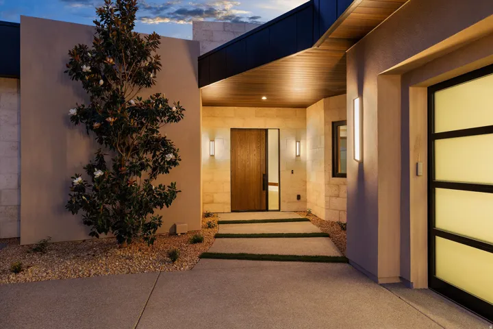 Modern entryway featuring a solid wood door with vertical pull handle, flanking wall sconces, natural stone block facade, and wood-finish soffit