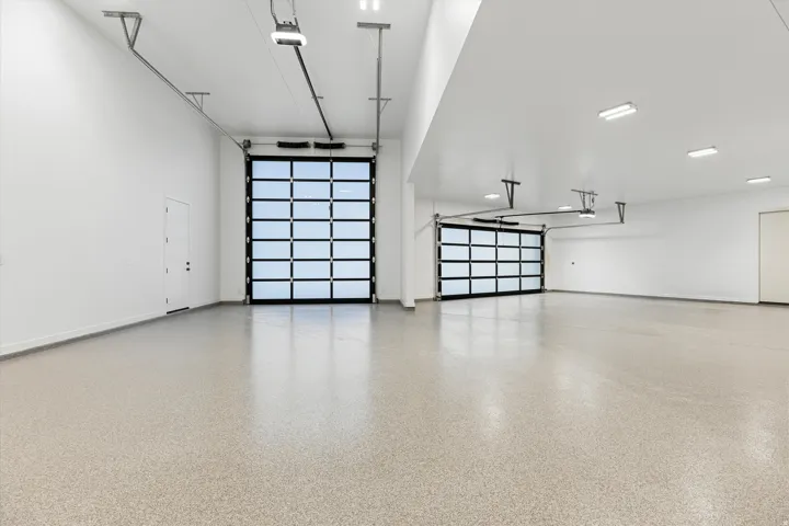 Large garage space featuring two glass-paneled overhead doors, a speckled epoxy floor finish, and multiple recessed and surface-mounted ceiling lights