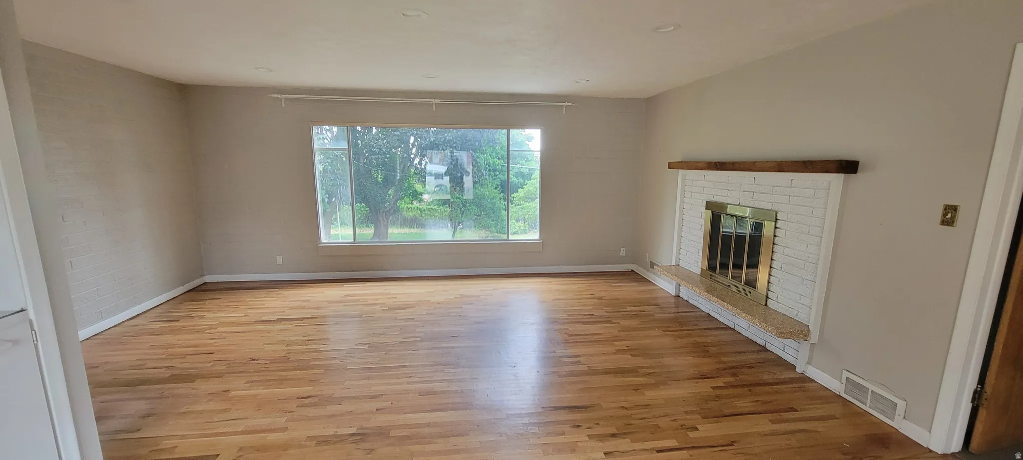 Spacious room featuring wood-finish flooring, a wide picture window, and a white brick fireplace with a wood mantle and brass-toned screen