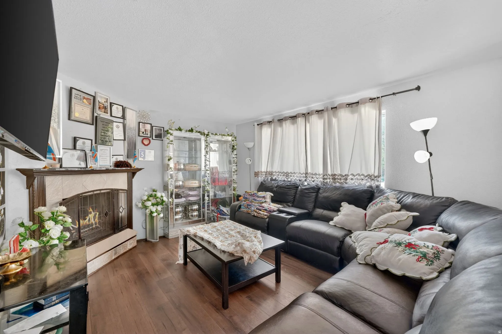 Living area featuring wood-finish flooring, a fireplace with a tiled surround and wood mantel, and expansive windows with privacy coverings