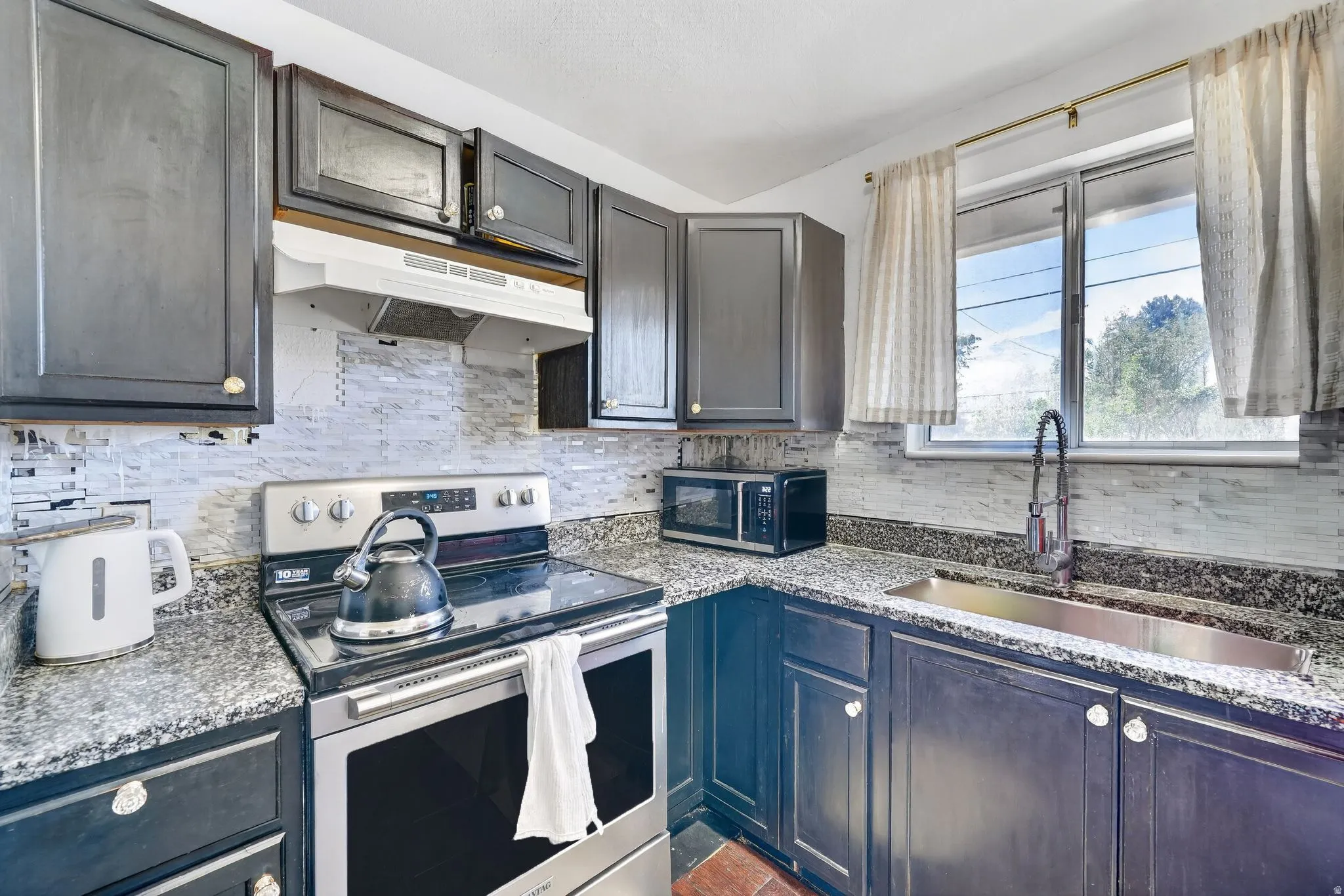 Kitchen featuring dark cabinetry, speckled countertops, a mosaic tile backsplash, stainless steel appliances, and a window with a pull-down faucet