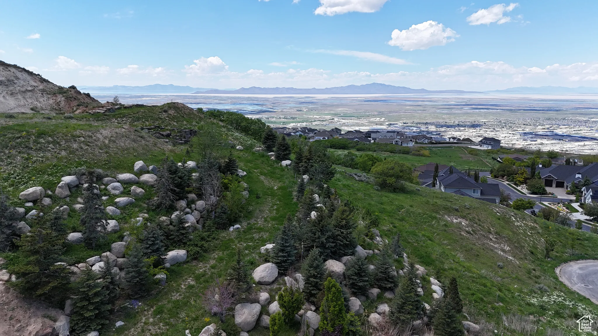 Elevated terrain featuring scattered boulders and evergreen trees with distant residential structures and a large body of water under a clear sky