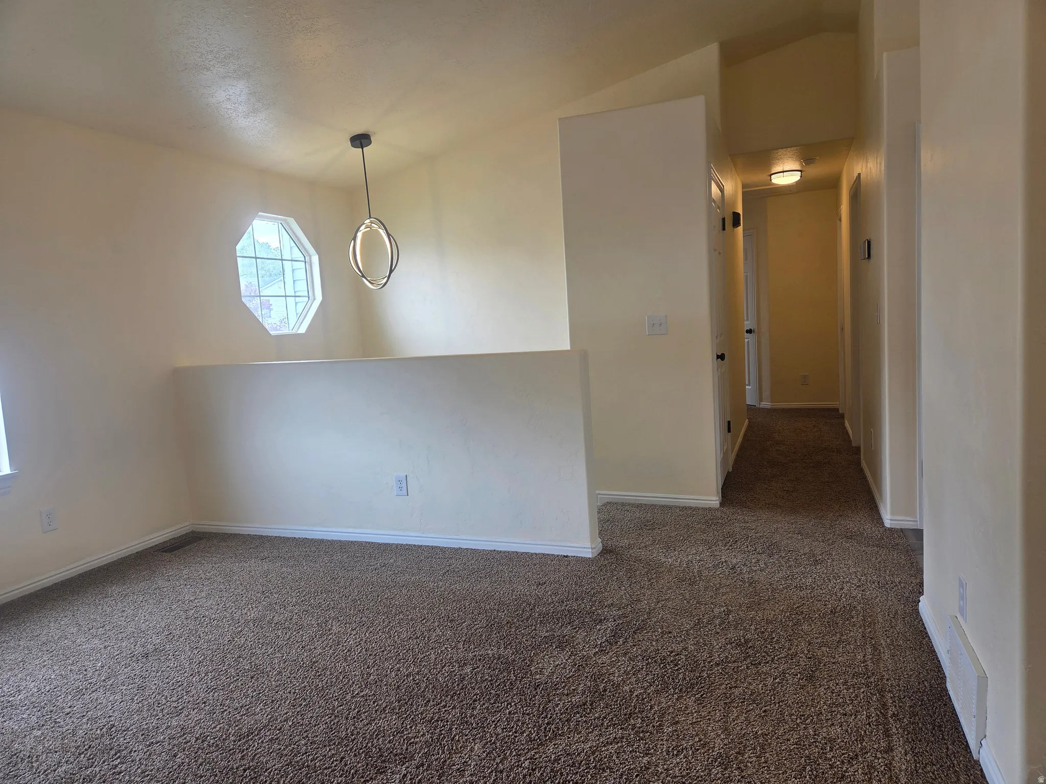 Carpeted interior space featuring a modern chandelier, octagonal window, vaulted ceiling, and a hallway with a flush-mount light fixture