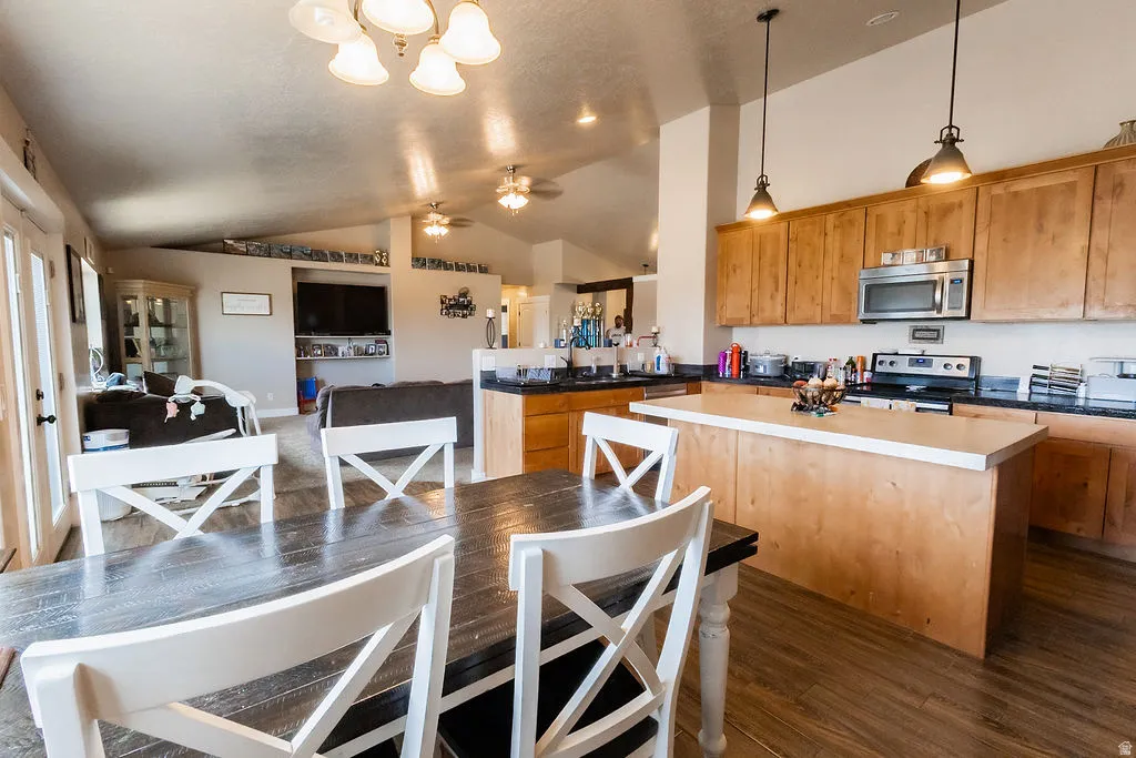 Open-concept living space featuring vaulted ceilings, wood-finish flooring, a kitchen with wood cabinetry and stainless steel appliances, and a central island with a light-colored countertop