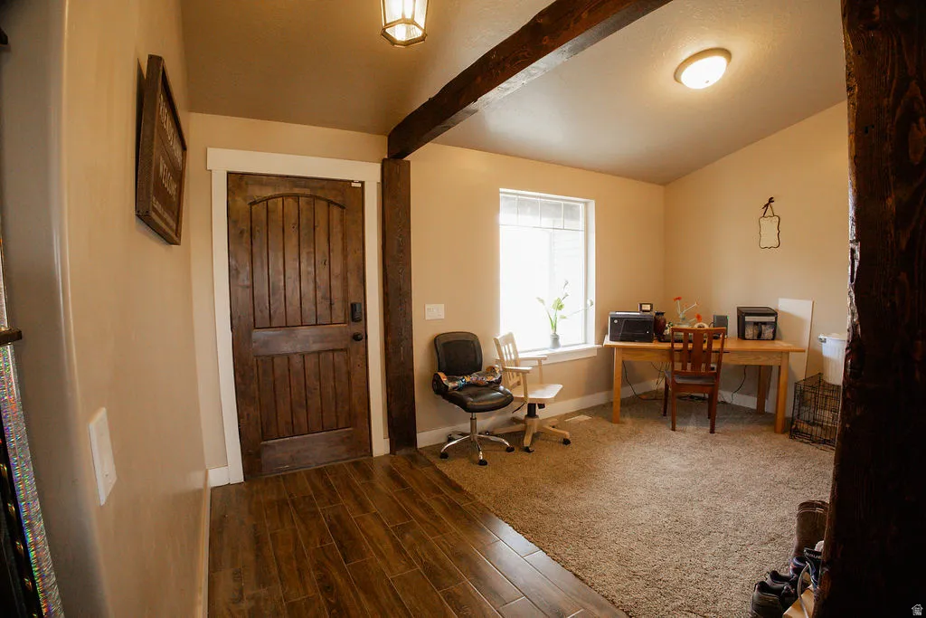 Entryway featuring wood-finish flooring transitioning to carpet, a paneled wood door with white trim, and exposed wood beam accents