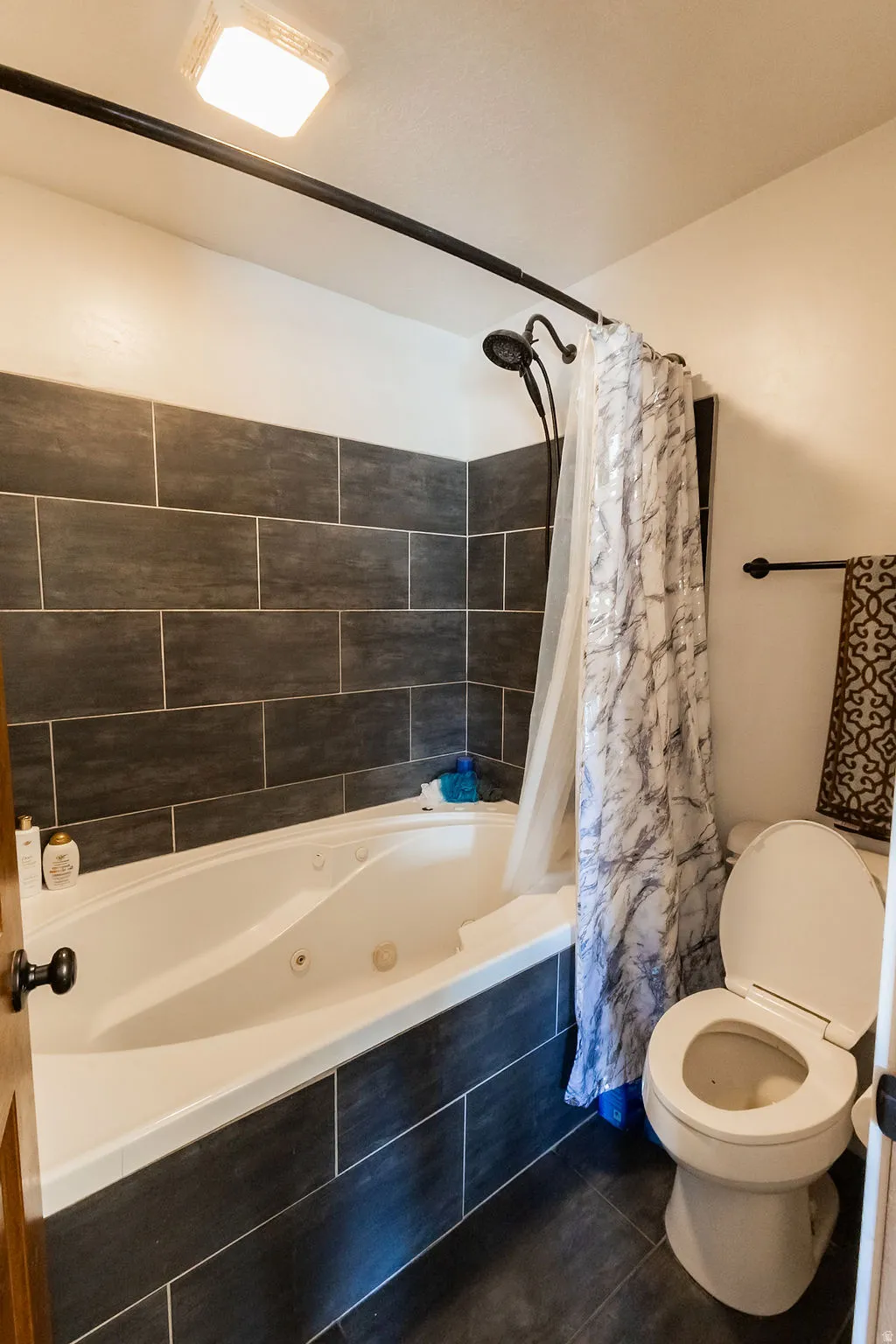 Bathroom featuring a built-in jetted tub with dark rectangular wall tiling, a coordinating dark tile floor, and a matte black shower fixture