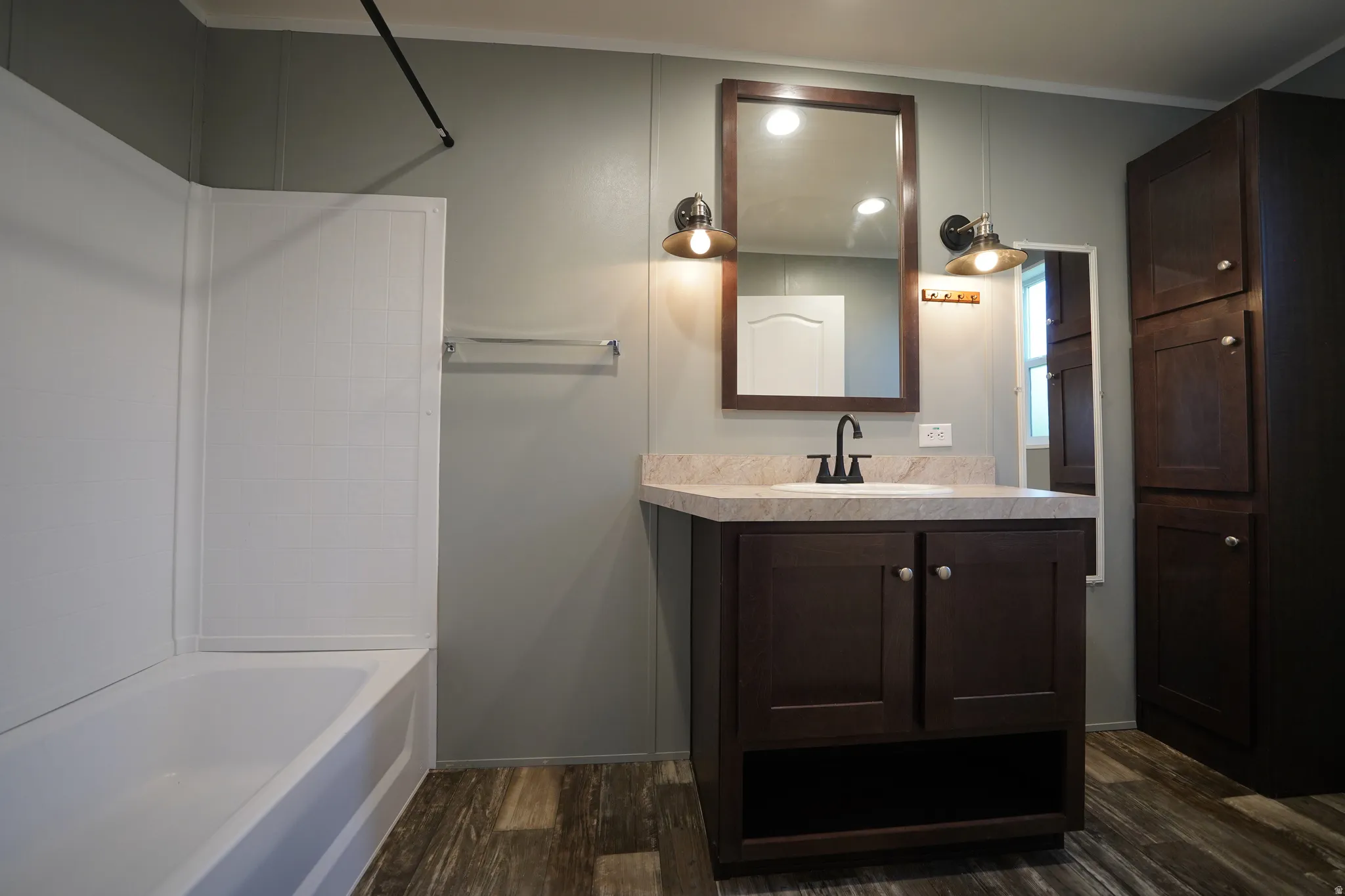 Primary Bathroom featuring wood-finish flooring, a white bathtub with shower surround, a dark wood-tone vanity with a light-colored countertop and integrated sink, a framed mirror, and dual wall-mounted light fixtures