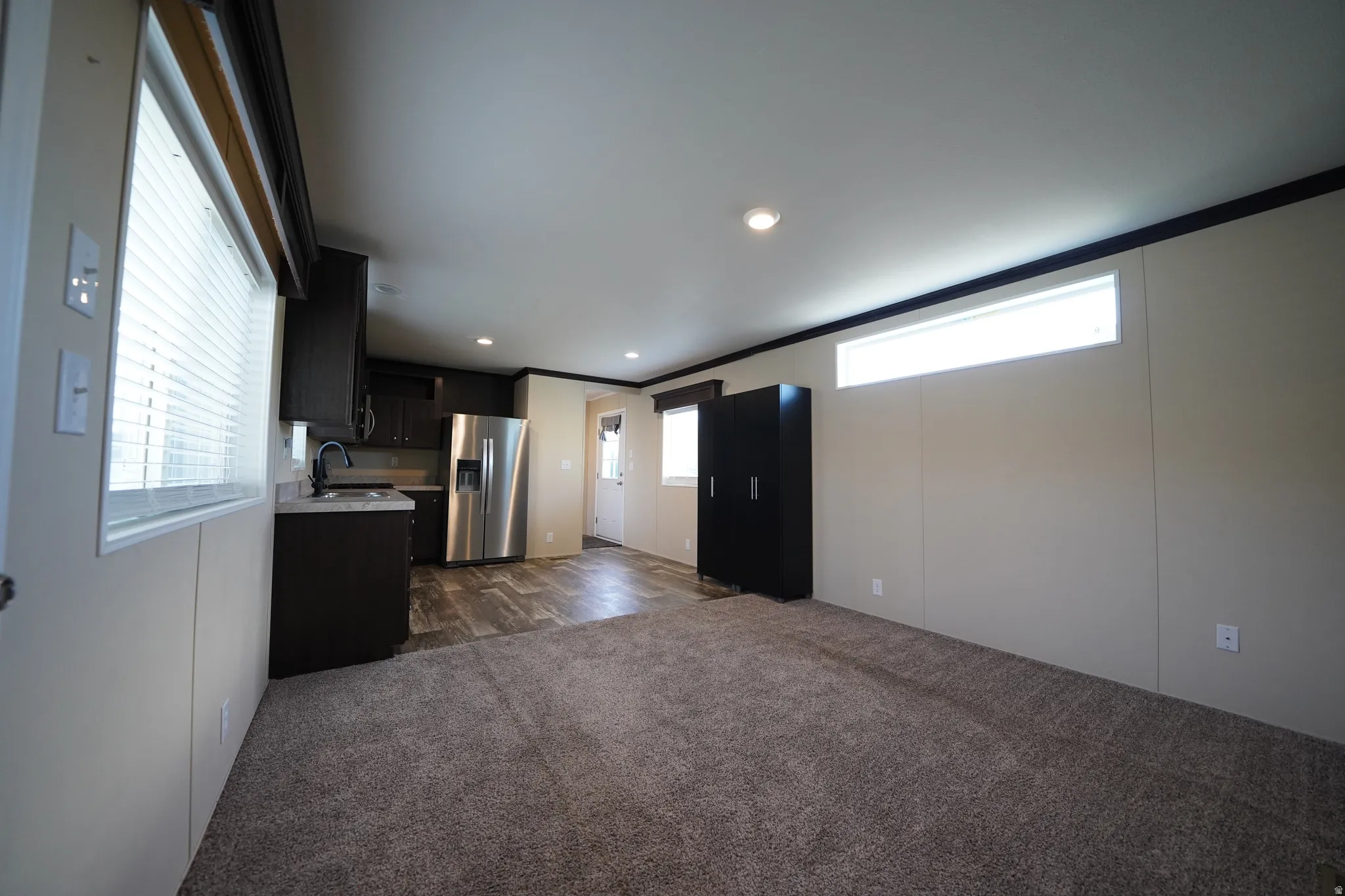 Open-concept interior featuring wood-finish flooring in the kitchen, stainless steel refrigerator, dark cabinetry, recessed lighting, and a neutral-toned carpeted area