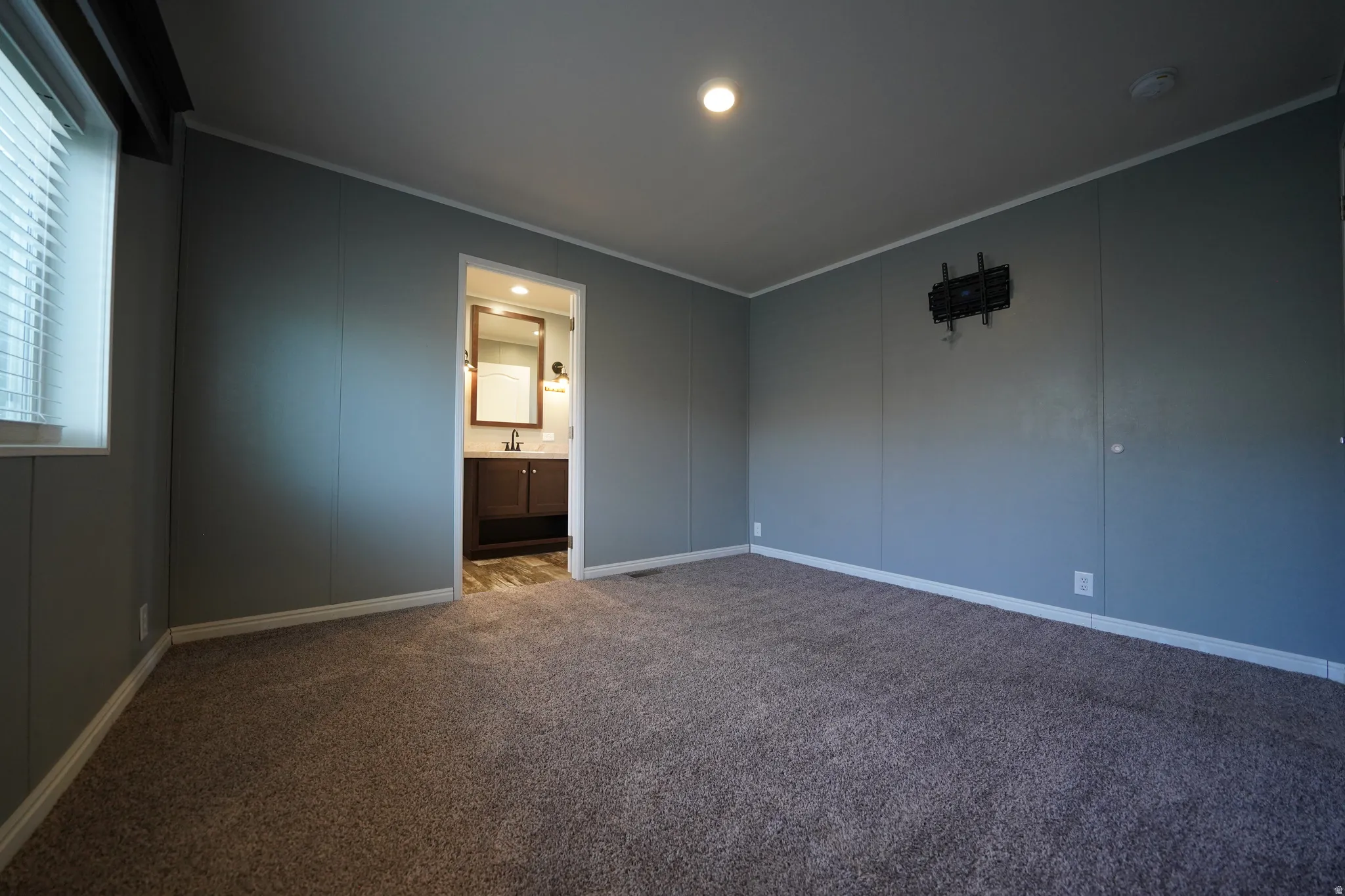 Primary Bedroom - Carpeted interior space with a window featuring blinds, an overhead light fixture, and a doorway revealing a bathroom with a vanity, mirror, and sconce lighting