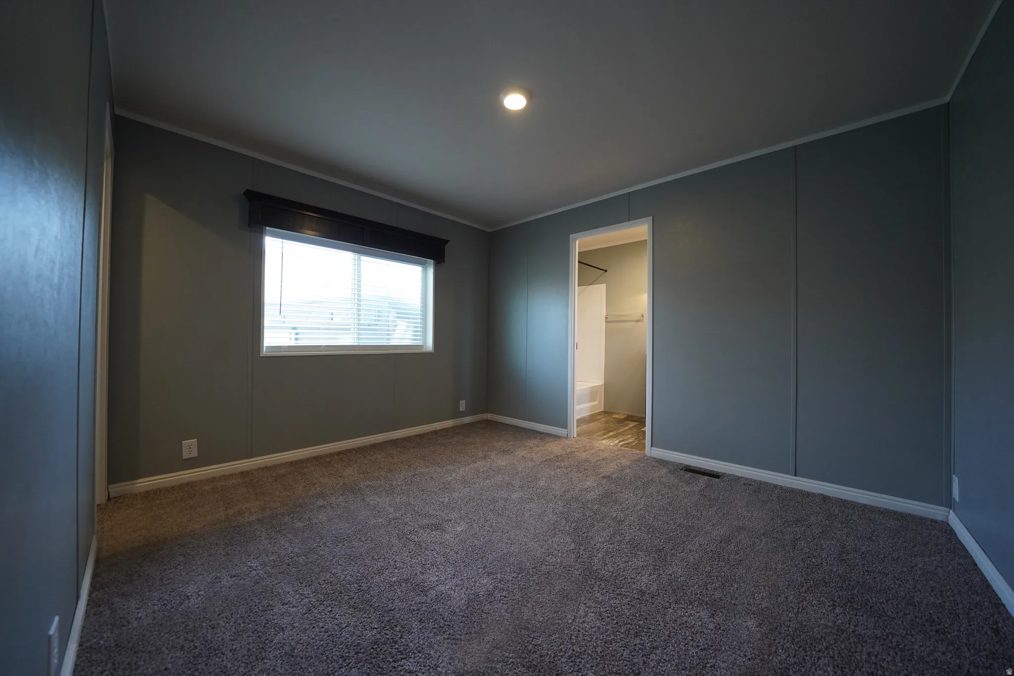 Primary Bedroom - Carpeted room featuring a window with blinds and a dark wood-finish valance, recessed ceiling lighting, and a doorway opening to a bathroom with a white bathtub and shower