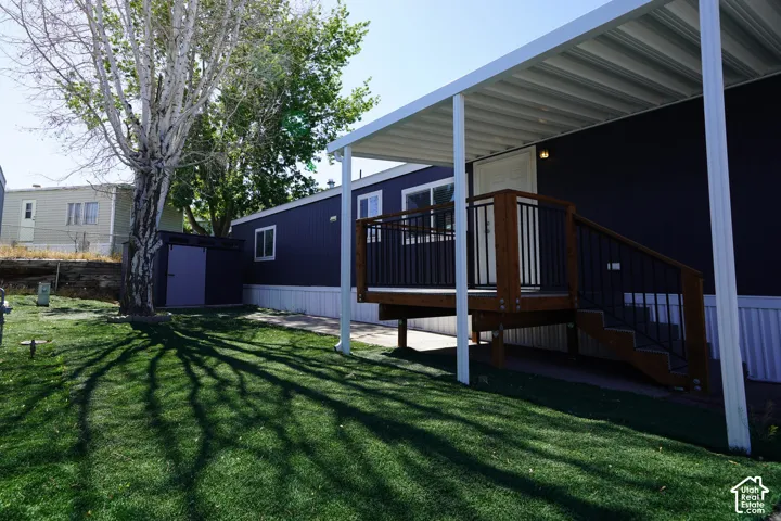 Manicured turf lawn with mature tree, accompanied by a dark blue siding structure featuring a covered porch with wooden deck and black metal railings, and a detached storage shed