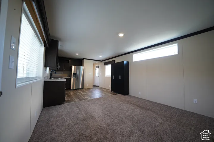 Open-concept interior featuring wood-finish flooring in the kitchen, stainless steel refrigerator, dark cabinetry, recessed lighting, and a neutral-toned carpeted area