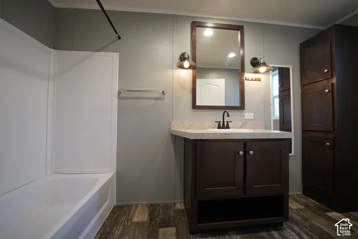 Primary Bathroom featuring wood-finish flooring, a white bathtub with shower surround, a dark wood-tone vanity with a light-colored countertop and integrated sink, a framed mirror, and dual wall-mounted light fixtures