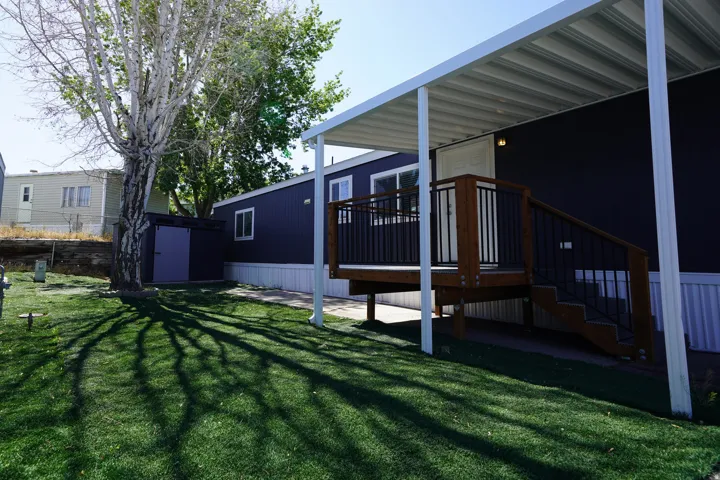Manicured turf lawn with mature tree, accompanied by a dark-sided structure featuring a covered porch with wooden deck and black metal railings, and a detached storage shed