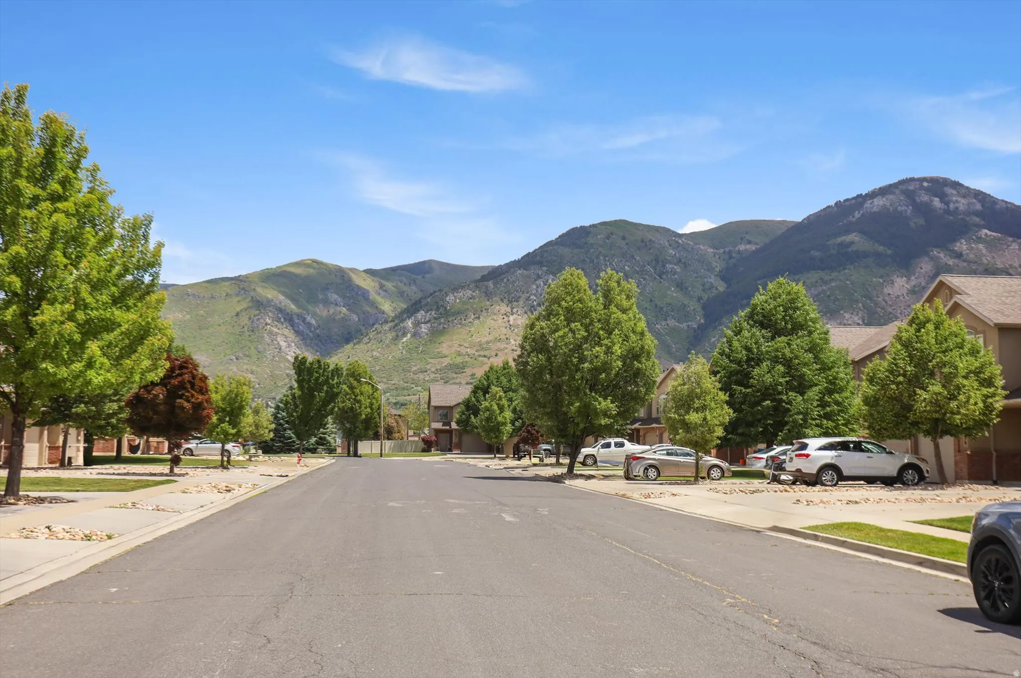 Neighborhood street featuring mature trees, paved sidewalks, and mountain scenery